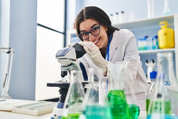 Young hispanic woman wearing scientist uniform using microscope at laboratory