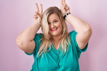 Fototapeta premium Caucasian plus size woman standing over pink background posing funny and crazy with fingers on head as bunny ears, smiling cheerful