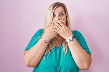 Caucasian plus size woman standing over pink background shocked covering mouth with hands for...