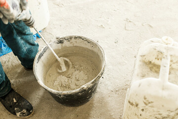 Worker mixing gypsum plaster with water for plastering walls. Construction of house and home renovation concept. Close up of bucket with putty mix and handyman hands