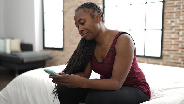 African woman using smartphone sitting on bed looking angry at bedroom - Powered by Adobe