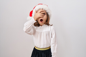 Little hispanic girl wearing christmas hat peeking in shock covering face and eyes with hand,...