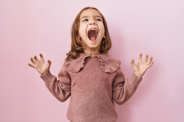 Little hispanic girl standing over pink background crazy and mad shouting and yelling with...