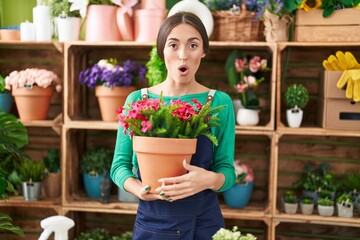 Young hispanic woman working at florist shop holding plant scared and amazed with open mouth for surprise, disbelief face