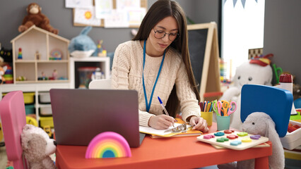 Young beautiful hispanic woman preschool teacher using laptop writing on document at kindergarten