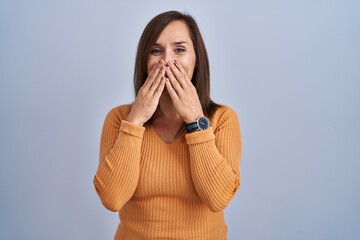 Middle age brunette woman standing wearing orange sweater laughing and embarrassed giggle covering...