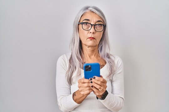 Middle age woman with grey hair using smartphone typing message relaxed with serious expression on face. simple and natural looking at the camera.
