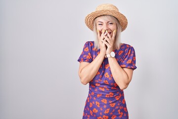 Young caucasian woman wearing flowers dress and summer hat laughing and embarrassed giggle covering...
