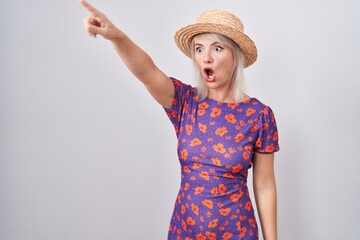 Young caucasian woman wearing flowers dress and summer hat pointing with finger surprised ahead, open mouth amazed expression, something on the front