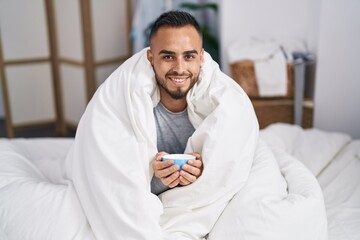 Young hispanic man drinking cup of coffee sitting on bed at bedroom