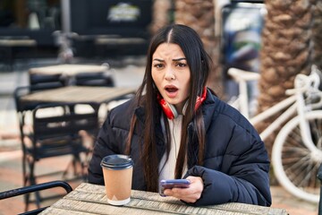 Young hispanic woman using smartphone sitting on the table scared and amazed with open mouth for...