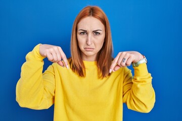 Young woman standing over blue background pointing down looking sad and upset, indicating direction with fingers, unhappy and depressed.