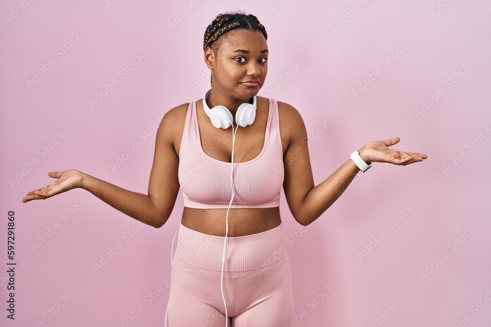 Poster African american woman with braids wearing sportswear and headphones clueless and confused expression with arms and hands raised. doubt concept.