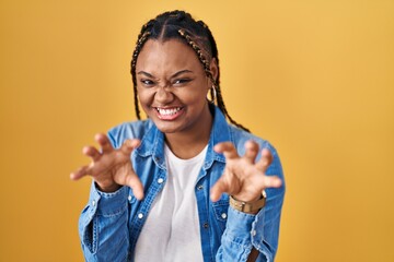 African american woman with braids standing over yellow background smiling funny doing claw gesture...