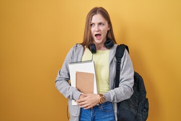 Young caucasian woman wearing student backpack and holding books angry and mad screaming frustrated and furious, shouting with anger. rage and aggressive concept.