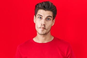 Obraz premium Young hispanic man standing over red background making fish face with lips, crazy and comical gesture. funny expression.