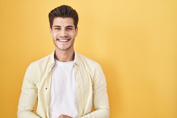 Young hispanic man standing over yellow background with hands together and crossed fingers smiling...