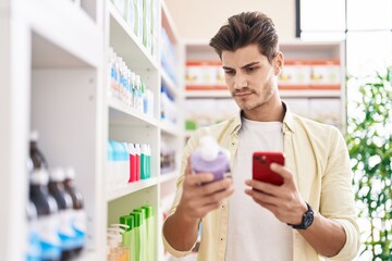 Young hispanic man customer using smartphone holding gel bottle at pharmacy