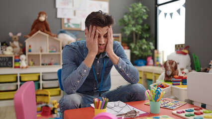 Young hispanic man preschool teacher stressed sitting on table at kindergarten