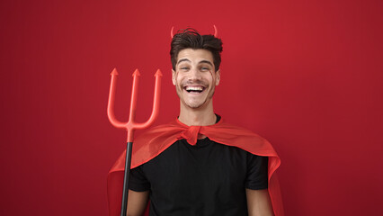 Young hispanic man wearing devil costume holding trident over isolated red background