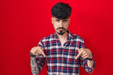 Young hispanic man with beard standing over red background pointing down looking sad and upset, indicating direction with fingers, unhappy and depressed.