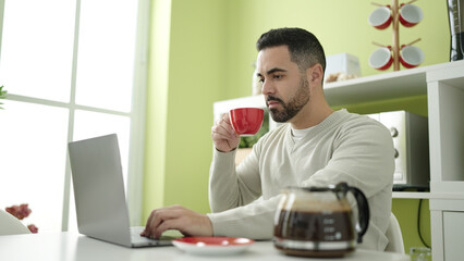 Young hispanic man using laptop drinking coffee at home