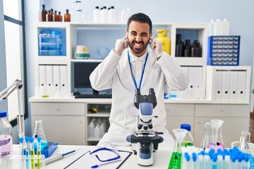 Young hispanic man with beard working at scientist laboratory covering ears with fingers with annoyed expression for the noise of loud music. deaf concept.