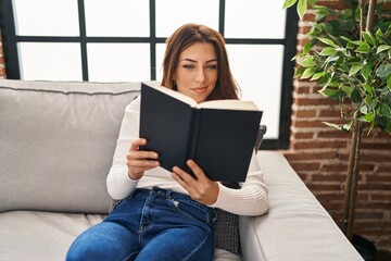 Young hispanic woman reading book sitting on sofa at home