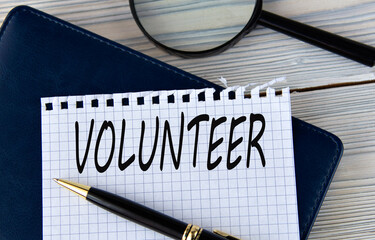 The word VOLUNTEER on a white sheet of paper against the background of a fragment of a notebook and a pen