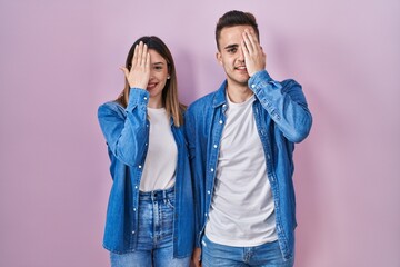 Young hispanic couple standing over pink background covering one eye with hand, confident smile on face and surprise emotion.