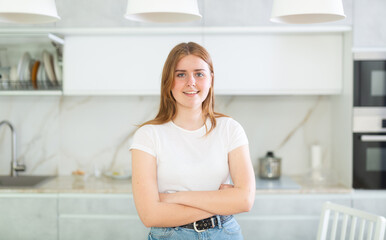 Portrait of positive girl teenager in casual wear smiling at camera while standing with crossed hands in cozy kitchen
