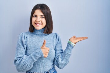 Young hispanic woman standing over blue background showing palm hand and doing ok gesture with...