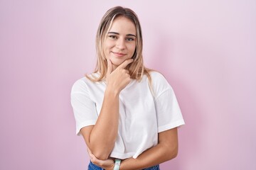 Young blonde woman standing over pink background looking confident at the camera smiling with crossed arms and hand raised on chin. thinking positive.