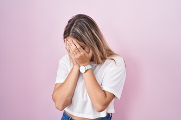 Young blonde woman standing over pink background with sad expression covering face with hands while crying. depression concept.