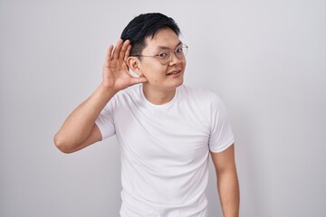 Young asian man standing over white background smiling with hand over ear listening an hearing to rumor or gossip. deafness concept.