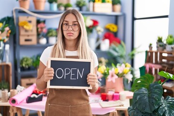 Young blonde woman working at florist holding open sign relaxed with serious expression on face. simple and natural looking at the camera.