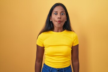 Young indian woman standing over yellow background making fish face with lips, crazy and comical gesture. funny expression.