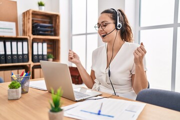 Young hispanic woman wearing call center agent headset at the office very happy and excited doing winner gesture with arms raised, smiling and screaming for success. celebration concept.
