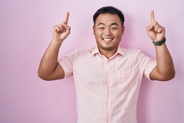 Chinese young man standing over pink background smiling amazed and surprised and pointing up with fingers and raised arms.