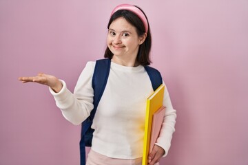 Woman with down syndrome wearing student backpack and holding books smiling cheerful presenting and pointing with palm of hand looking at the camera.