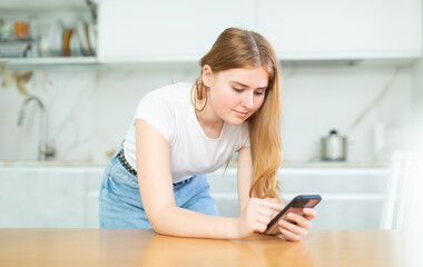 bored young girl stands in kitchen and holds smartphone in hands. woman reads news and likes photos of friends on social networks
