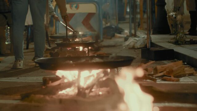 Slow motion footage from a low-angle view of paellas cooking on an open flame on a fenced street during the Fallas festival in Valencia, Spain