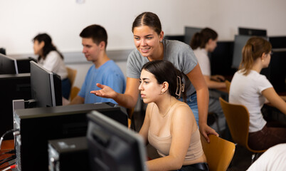 Positive female student explaining the topic to friend while she is studying computer science on the PC in the classroom.