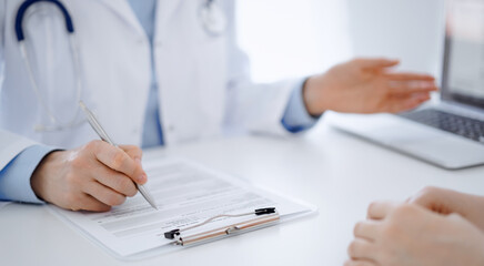 Doctor and patient sitting opposite each other at the desk in clinic. The focus is on female physician's hands filling up the medication history record form or checklist, close up. Medicine concept