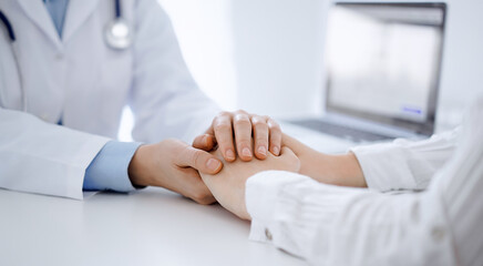Doctor and patient sitting opposite each other at the table in clinic office. The focus is on female physician's hands reassuring woman, only hands, close up. Medicine concept - 597307456