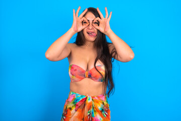 beautiful brunette woman wearing bikini over blue background doing ok gesture like binoculars sticking tongue out, eyes looking through fingers. Crazy expression.