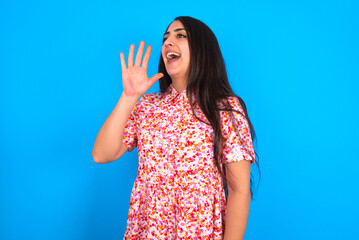 Fototapeta premium beautiful brunette woman wearing floral dress over blue background look empty space holding hand face and screaming or calling someone.