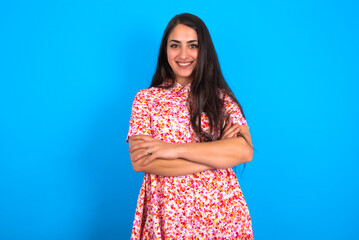 Confident beautiful brunette woman wearing floral dress over blue background with arms crossed...