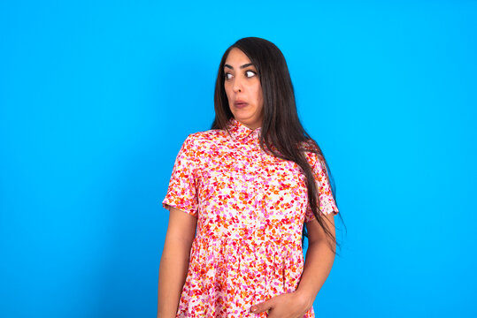 Shocked beautiful brunette woman wearing floral dress over blue background look empty space with open mouth screaming: Oh My God! I can't believe this.
