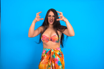 beautiful brunette woman wearing swimwear over blue background Shouting frustrated with rage, hands trying to strangle, yelling mad.